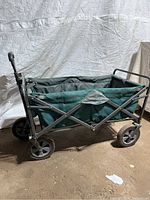 Side view of the foldable beach cart showing the green fabric, metal frame, and plastic wheels.