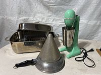 Lot overview showing green milkshake mixer with stainless steel mixing cup, stainless steel roasting pan with lid, and metal cone strainer with black handle.