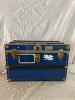 Front view of a large blue metal vintage trunk with brass corner protectors, latches, and lock, showing wear and scratches.