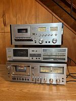 Front view of three vintage cassette decks stacked on a table in front of a wood-paneled wall, showing controls, tape compartments and meters.