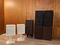 Lot of 7 speakers arranged showing two white pedestal-mounted speakers, one medium dark brown speaker, and four large brown speakers stacked in pairs. Wood-paneled wall background with 5ft tape measure for scale.