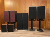 Photo showing entire group of eight speakers with various sizes and styles against a wood-paneled wall with a tape measure next to them for scale (5ft).