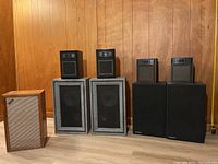 Wide shot of nine speakers lined up against wood panel wall with tape measure at 5 feet showing scale.