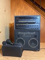Front view of MegaStar HP-K8000 karaoke machine featuring dual round speakers and control knobs. Two black wired microphones and a zippered carrying case in front on wooden floor.