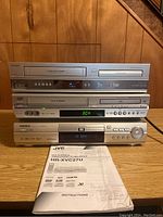 Stacked view of three silver media players on wooden surface with front panles visible showing disc trays, buttons, and displays. JVC instruction manual placed in front.