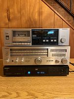 Full frontal view of stacked Sanyo RD220 stereo cassette deck (top), Sony STR-333 FM-AM receiver (middle), and Sony DVP-NS300 DVD player (bottom) on wooden surface under staircase