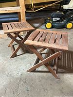 Two foldable teak stools shown side by side on floor; both have slatted rectangular tops and foldable X-leg bases