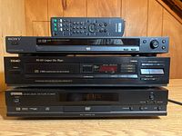 Photo showing all three players stacked: Sony DVP-NS325 with remote on top, Teac PD-365 in middle, Yamaha DV-S5450 on bottom on a wooden surface