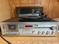 Front view of stereo with vinyl turntable, cassette deck, 8-track player, LED clock, and AM/FM tuner. Woodgrain cabinet and protective dust cover visible.