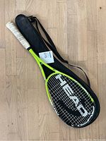 Full view of the Head tennis racket with white grip tape and black carrying case on wood floor