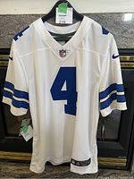 Front view of the white Dallas Cowboys jersey with number 4 and blue stripes on the sleeves hung on a hanger.