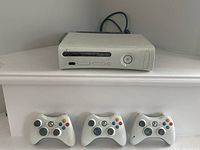 Xbox 360 console with three white wireless controllers lined in front. Console shows signs of use with slight discoloration. Two controllers missing battery covers.