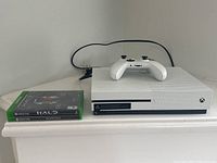 Xbox One S console with wireless controller and two game cases for Halo: The Master Chief Collection and Injustice 2 shown from front angle.