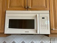 Front view of the clean white Samsung over-the-range microwave mounted under wooden cabinets with control panel and handle visible.