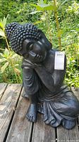 Frontal view of the black resting Buddha statue seated outdoors on a wooden surface with greenery in the background.