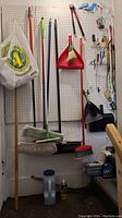 Cleaning tools hanging on a pegboard including brooms, mops, dustpans, hand tools, and bags.