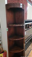Front view of dark wood corner shelf cabinet with four curved shelves including one with brass railing.