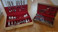 Two sets of flatware in open canteens, one cream-colored with silverplate utensils, one wooden with rosewood-handled utensils and serving knives, arranged for display.