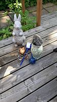 Group of garden decor items on wooden deck, includes rabbit statue, mosaic vase, two glass orbs and frog figurine.