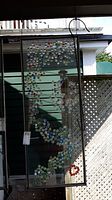 Full vertical shot of framed glass art panel showing colorful glass marbles arranged in swirling pattern. The glass is clear and the marbles are blue, red, clear, and green.
