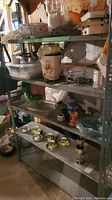 Gray metal shelving unit with various household items such as glassware, ceramics, and decorative items on shelves. Basement setting.