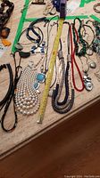 Photo showing sixteen assorted necklaces laid on a table with a tape measure for scale, featuring faux pearl strands, multi-colored beads, and pendants.
