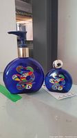 Two cobalt blue glass bottles with colorful snake-like abstract designs on a white surface. One bottle has a silver spherical cap, the other a gold collar pump dispenser.