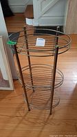 Angle view of brass plant stand showing three oval wire shelves and four vertical legs on hardwood floor.