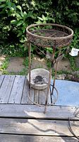 Full view of the metal plant stand with decorative circular top and base, showing weathered condition and outdoor placement.
