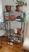 Shelving unit shown with various potted plants and decorative items on the glass shelves and top, against a light wall and beside chairs.
