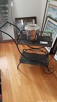 Photo of the black metal tea trolley with two wicker shelves and metal ice bucket on top, showing the full item from a side angle on a wood floor with framed pictures in background.