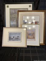 Four framed Canadiana artwork prints, arranged leaning against a dark surface. The prints depict scenes including loons on water and an autumn forest.