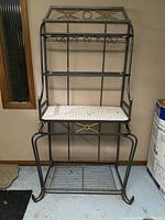 Front view of the baker's rack showing the metal frame, white marble-style middle shelf, built-in wine glass holder, and metal wire bottom shelf.