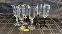 Seven fluted glasses displayed on a patterned cloth surface. Five with clear stems and two with black stems.