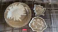 Three glass dishes on a dark cloth background showing holiday etched designs: one round with pine cones, one tree-shaped, and one scalloped with carolers.