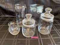 Five glass storage canisters arranged on cloth background showing front view with lids on some jars