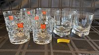 Set of 8 glass tumblers arranged in two rows showing four RCR crystal glasses on left with distinctive leaf pattern and stickers, and four plain faceted glasses on right.