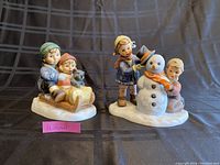 Two Christmas-themed Hummel Goebel figurines together on a black grid cloth background