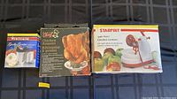 Photo showing boxed garlic peeler/dicer, boxed chicken roaster, and boxed apple peeler on blue plaid background.