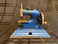 Side view of the blue miniature hand crank sewing machine showing the needle area, sewing platform, crank wheel with wooden handle, and gold decoration on metal body.