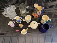 Wide view showing all mugs and jugs including a tall floral jug, clear glass beer mug, and cut crystal jug with base damage.