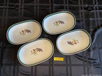 Photo shows 4 oval dishes arranged on a black cloth with a central fruit design and green and gold borders.