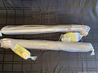 Two metal roller shades sealed in plastic packaging with hardware attached, lying horizontally on a dark fabric surface.