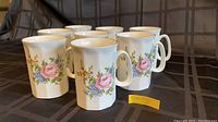 Front view of eight white ceramic mugs with pink rose and floral designs, arranged in two rows.