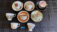 Eleven assorted vintage tea cups arranged on a table, seven of which have corresponding saucers. The cups feature a variety of floral patterns, gold trim, and different cup shapes including footed and traditional styles.