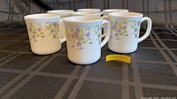 Front angle showing 7 out of 8 mugs with violet floral pattern on white ceramic.