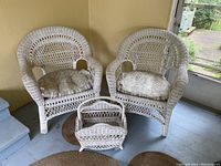 Two white wicker armchairs with patterned cushions and a matching magazine rack placed on the floor beside them.