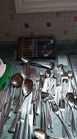 Overview of assorted cutlery items including souvenir spoons, forks, knives, and large serving spoons laid on a metal surface