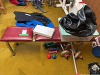 Weight lifting bench with dumbbells underneath, and various exercise accessories on top including a black seat cover and plastic container.