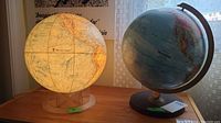 Photo showing two globes side by side on a table near a window with curtains, one lighted and one not.